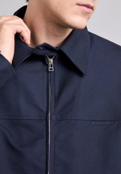 Navy blue jacket with a zip front, pointed collar, and subtle branding. Features solid fabric and a tailored fit.