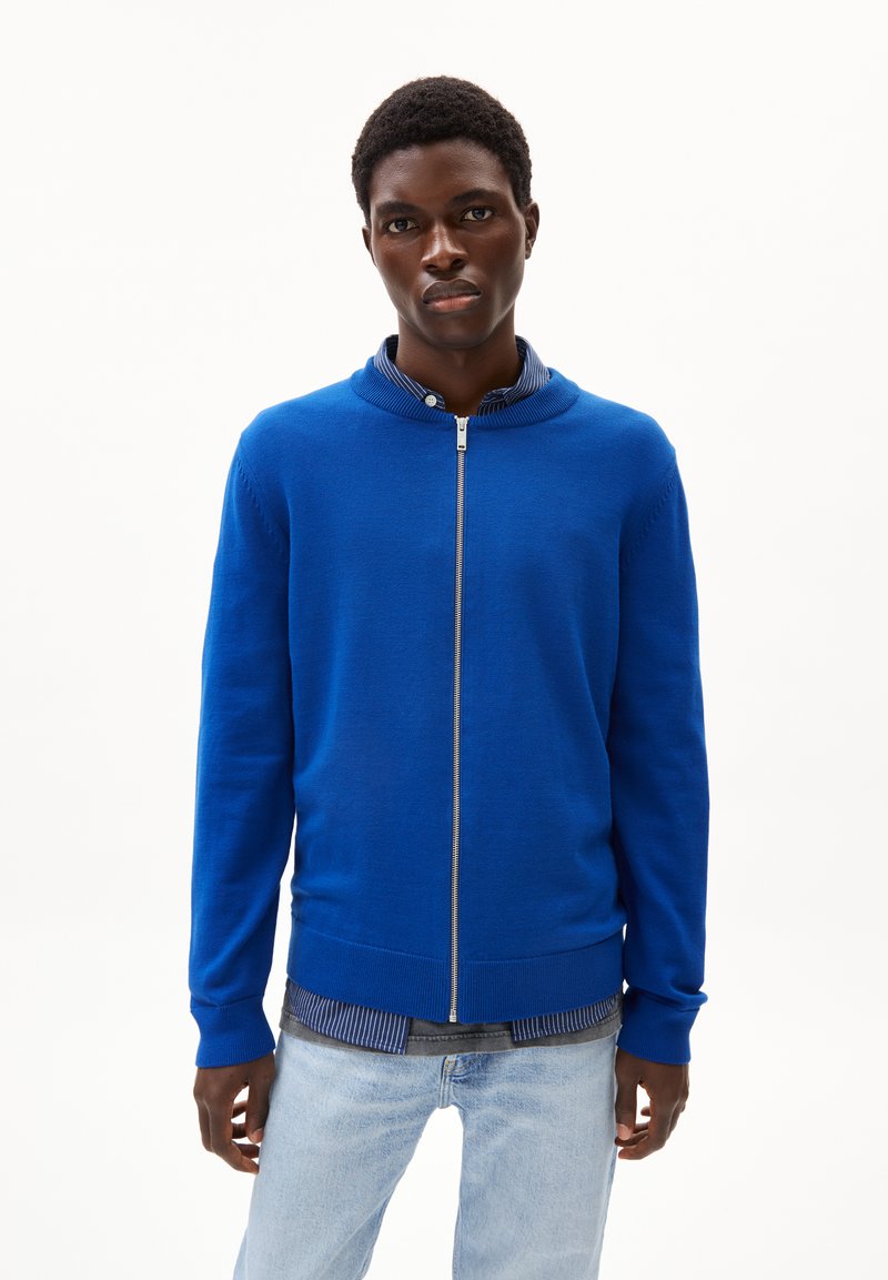 Blaues Zip-Up-Sweatshirt mit rundem Kragen, gerippten Bündchen und glatter Textur, über einem gestreiften Hemd getragen und mit heller Jeans kombiniert.