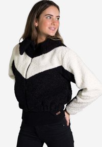 Chaqueta de forro polar en blanco y negro con diseño en forma de chevron, cierre con cremallera, cuello puntiagudo y cinturilla de punto negra. Tejido texturizado con un acabado suave.