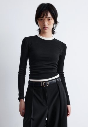 Sort langærmet top med hvid krave trim, lavet af ribbet stof. Kombineret med høj talje sorte bukser og et smalt sort bælte.