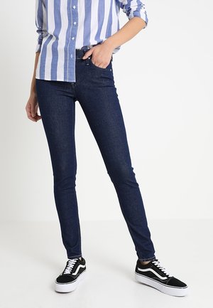 Jeans skinny azul oscuro con una textura suave, combinados con una camisa de rayas azul claro y blanca. El calzado son zapatillas negras con detalles blancos.