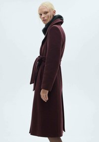 Mørkerød uldfrakke med bælte i taljen, lange ærmer og krave foret med sort pels, med en lige silhuet og sideskår.