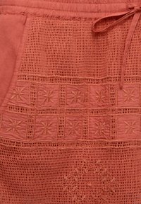 Vêtement couleur rouille avec des motifs crochetés complexes et des motifs floraux brodés, doté d'un tissu texturé et d'une taille réglable par cordon.