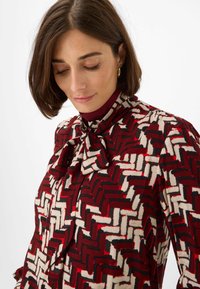 Long-sleeve blouse with a high collar, featuring a red, black, and cream geometric pattern. Tied bow detail at neckline and button closure.