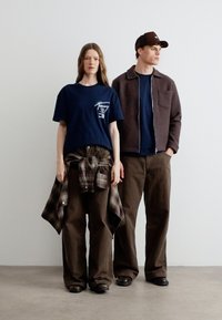 Δύο άτομα στέκονται δίπλα σε έναν απλό τοίχο, φορώντας καθημερινά σκούρα μπλε και καφέ ρούχα με φαρδιά παντελόνια και πολυεπίπεδα τοπ, το ένα με καρό πουκάμισο δεμένο γύρω από τη μέση.