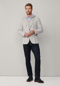 Grå stickad blazer med huva, enkel knappstängning, två framfickor, lager över en ljusblå skjorta, i kombination med mörka jeans och bruna skor.