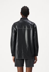 Giacca camicia in finta pelle nera con collo, cuciture bianche e polsini con bottoni, abbinata a pantaloni corti in denim scuro.