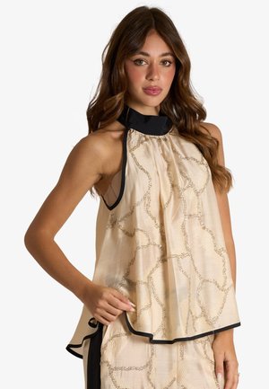 Giovane donna con lunghi capelli mossi che indossa una canotta beige senza maniche con bordi neri e motivo astratto, posando con una mano sul fianco.