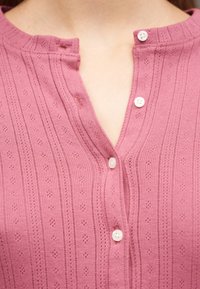 Gros plan sur un haut en maille rose texturée avec de petits boutons blancs et un décolleté partiellement ouvert.