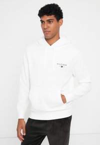 Hoodie branco feito de algodão, com um bolso frontal, capuz ajustável por cordão e um pequeno logótipo no peito. Textura suave com punhos canelados.