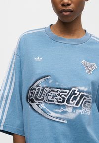 Άτομο που φορά ένα μπλε φαρδύ μπλουζάκι Adidas με λευκές ρίγες στα μανίκια και το γραφικό κείμενο "questra" μπροστά.