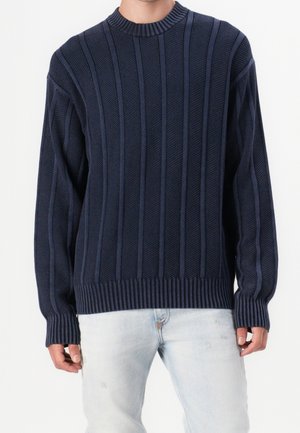 Homme portant un pull en maille côtelée bleu marine avec des rayures texturées verticales et un jean bleu clair déchiré, debout devant un fond uni.