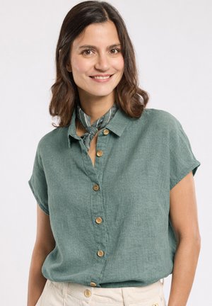 Femme souriante aux cheveux bruns mi-longs portant une chemise verte à manches courtes boutonnée, un pantalon beige et un foulard à motifs autour du cou.