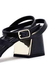 Sandalias de tacón negras de cuero con un chunky tacón bloque metálico dorado, que cuentan con delgadas correas ajustables y un diseño de punta cuadrada.