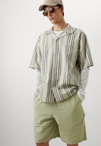Camisa de manga corta a rayas verdes y beiges con botones, combinada con pantalones cortos texturizados de color verde claro. Los accesorios incluyen gafas de sol y una gorra.