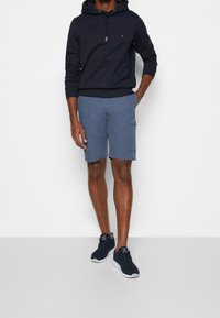 Marineblauer Hoodie mit Kordelzug, blaue Cargo-Shorts mit Seitentaschen und schwarze Sneakers mit weißen Sohlen. Einfaches und funktionelles Outfit.