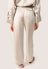 Beige vida byxor i slätt tyg, med en bakficka och eleganta sömmar. Matchande med svarta slidesandaler.