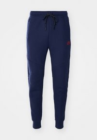 Marinblå Nike joggers med avsmalnande ben, justerbart snörliv, sidofickor och röd logotyp på låret mot vit bakgrund.