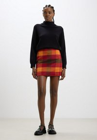 Maglione nero in maglia con collo alto, abbinato a una mini gonna a quadri rossi, arancioni e gialli. La modella indossa scarpe nere lucide.