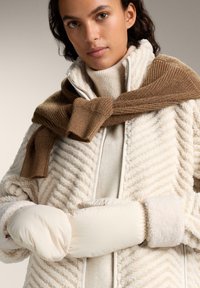 Krem-kolora lažno krzno jakna sa zigzag teksturom, složena preko bež pletene dukserice, uz bež rukavice i smeđu pletenu maramu.