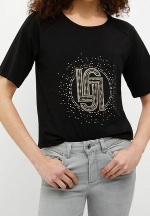 Camicia nera a maniche corte con motivo circolare di piccole borchie e lettere stilizzate “L” e “G” sul davanti, indossata con jeans grigio chiaro.