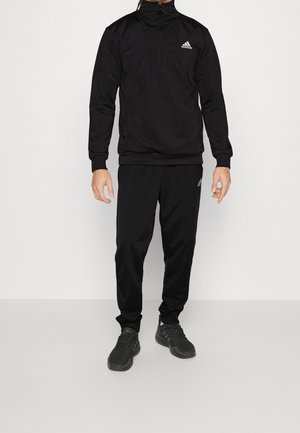 Mężczyzna w czarnym dresie Adidas z półgolfem na zamek i pasujących czarnych butach sportowych, stojący na jednolitym jasnym tle.