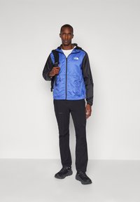 Giacca leggera blu e nera con cappuccio, zip e logo. Indossata sopra una maglietta bianca e pantaloni neri, accompagnata da uno zaino nero.