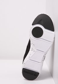 Chaussure de sport noire avec une semelle blanche texturée ornée de tâches noires, des accents en caoutchouc et un logo en relief sur le talon.
