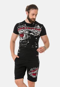 Schwarzes Baumwoll-T-Shirt mit weißem und rotem Grafikdruck, ausgestattet mit Spritzdetails. Kombiniert mit schwarzen Shorts, die ebenfalls grafische Akzente aufweisen.