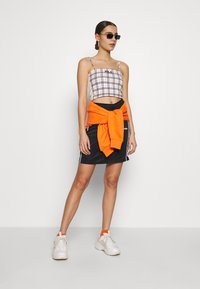 Top corto a quadri con spalline sottili, gonna nera con felpa arancione legata in vita e sneakers bianche con accenti arancioni.