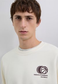 Krämfärgad sweatshirt med ribbad halsringning, med en mörkbrun tryckt logotyp med en basketboll och ränder. Mjuk, avslappnad tygstruktur.