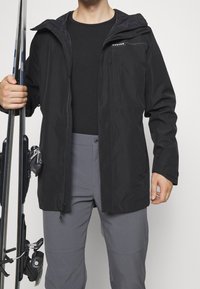 Czarna wodoodporna kurtka z zamkiem z przodu, regulowanym kapturem i logo na piersi. Model trzyma kije narciarskie, ubrany w szare spodnie.