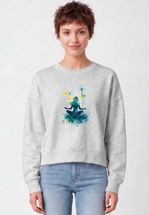 Jeune femme aux cheveux courts portant un sweat-shirt gris clair orné d'une figure de yoga colorée et de fleurs, associé à un jean bleu clair.