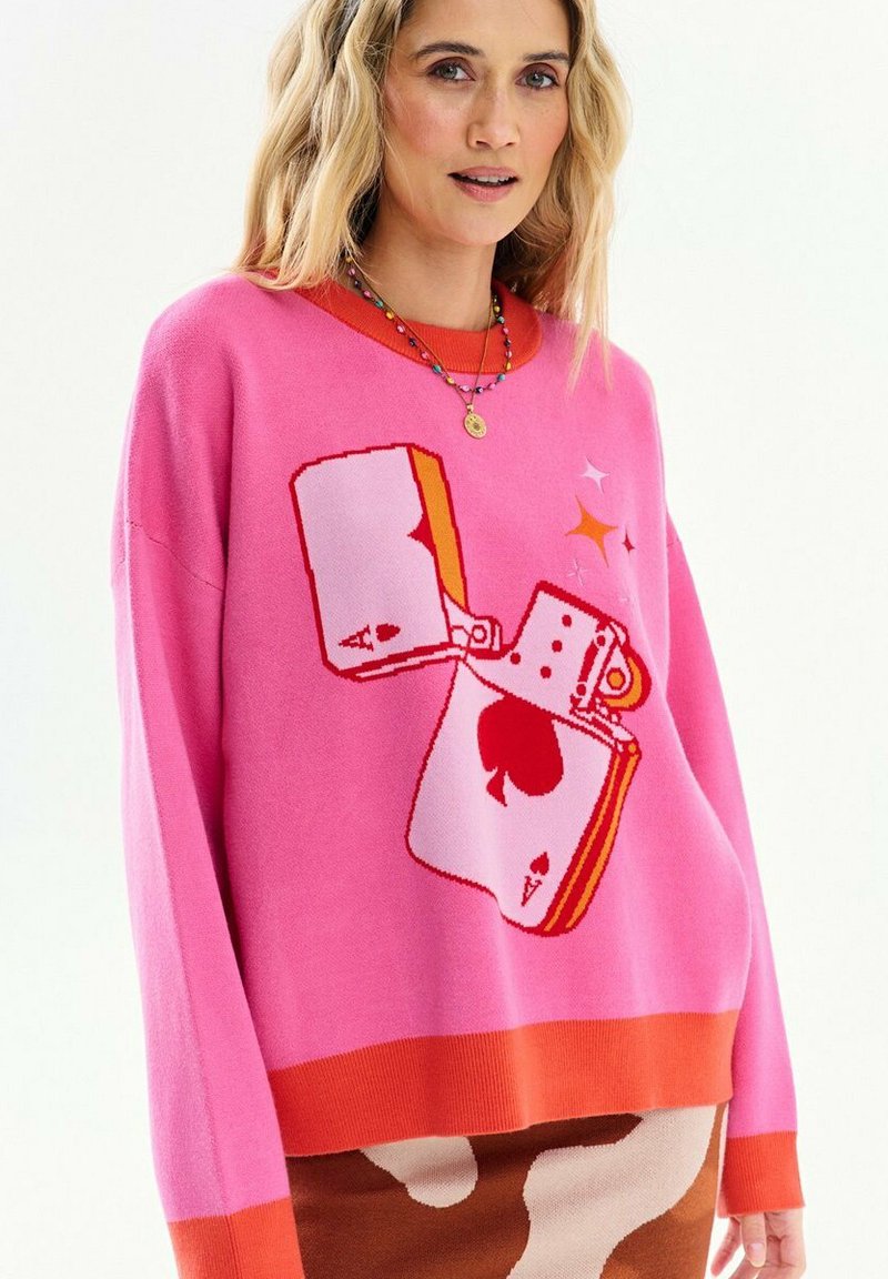 Frau trägt einen rosa-orangefarbenen Pullover mit einem Motiv einer brennenden Pik-Ass-Karte, kombiniert mit einem braun-cremefarbenen gemusterten Rock.