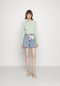 Camisola de manga longa em verde menta com gola, calções de ganga azul claros com um cinto estampado e sandálias de plataforma beges com alças verdes.