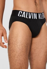Zwarte slip van zacht katoen, met een brede elastische tailleband met het "CALVIN KLEIN" logo in het wit. Goed gedefinieerde voorkant en beenopeningen.