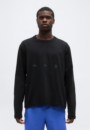 Hombre de pie, mirando al frente, vestido con una camiseta negra de manga larga y pantalones cortos azules, sobre un fondo blanco liso.