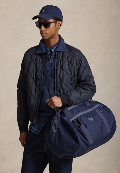 Άνδρας με μπλε σκουφί, γυαλιά ηλίου, μπουφάν με επένδυση και κρατώντας μεγάλο μπλε σακ βουαγιάζ στον ώμο, μπροστά σε ουδέτερο φόντο.