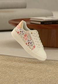 Λευκό αθλητικό παπούτσι με ροζ και μωβ ανθικό σχέδιο, μπεζ λεπτομέρεια στη φτέρνα και λευκά κορδόνια, αιωρούμενο πάνω από υφασμένο μπεζ χαλί κοντά σε ξύλινο τραπέζι.