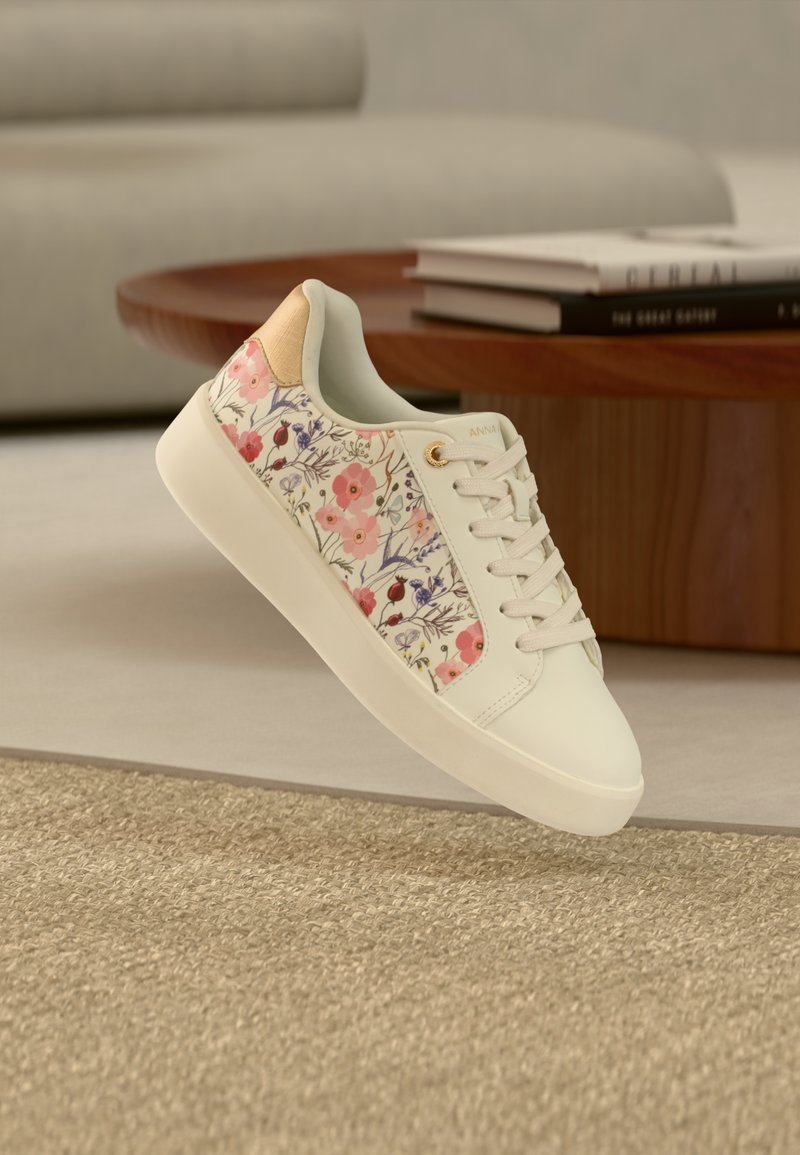 Λευκό αθλητικό παπούτσι με ροζ και μωβ ανθικό σχέδιο, μπεζ λεπτομέρεια στη φτέρνα και λευκά κορδόνια, αιωρούμενο πάνω από υφασμένο μπεζ χαλί κοντά σε ξύλινο τραπέζι.