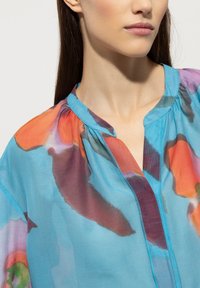 Leichte, transparente Bluse mit abstrakten Blumenmustern in Blau, Orange und Lila. Sie hat einen einfachen Kragen und Raffungselemente an den Schultern.