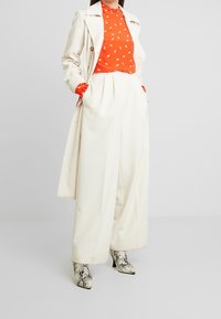 Witte trenchcoat over een oranje top met witte patronen, brede witte broek en enkellaarsjes met slangprint.
