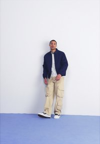 Jachetă marină, tricou gri, pantaloni cargo bej și adidași alb-negru; stând lângă un perete alb pe un covor albastru.