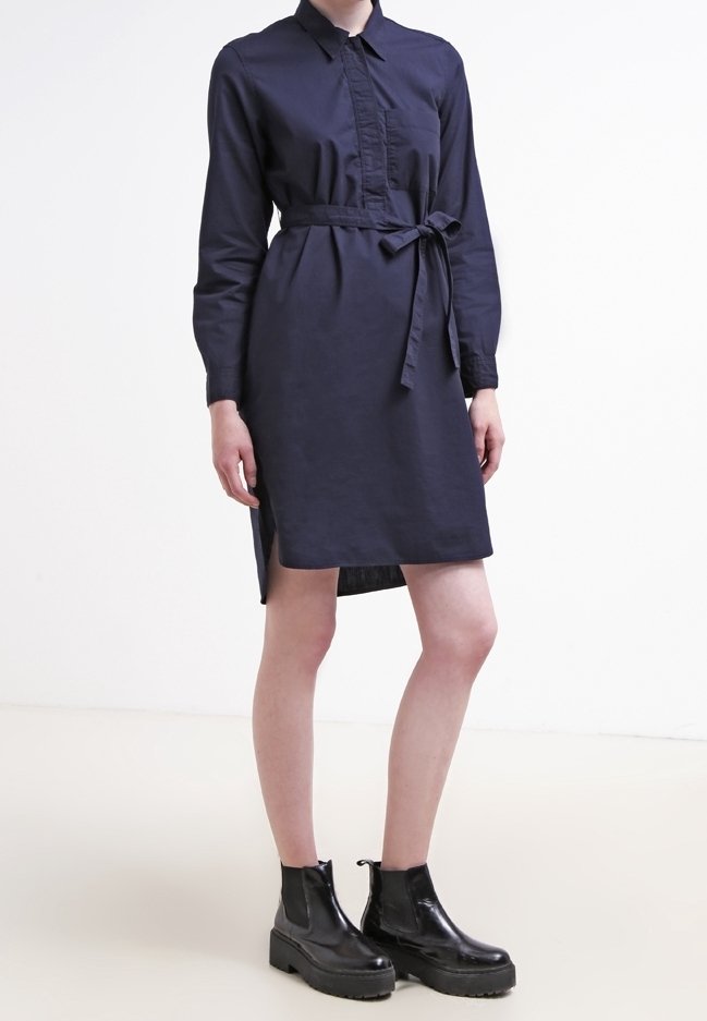 Femme portant une robe chemise bleu marine au genou avec ceinture à nouer et manches longues, associée à des bottines noires épaisses.