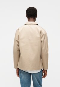 Μπεζ, αδιάβροχο μπουφάν με πίσω πάσχλο, απλό γιακά και ευθεία γραμμή. Συνδυασμένο με λευκό πουκάμισο με ρίγες και μπλε τζιν.