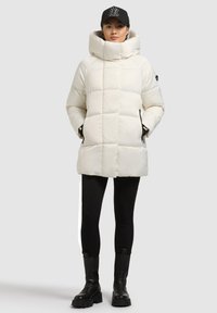 Biely prešívaný kabát s vysokým golierom, s prešívaným vzorom a bočnými vreckami, v kombinácii s čiernymi legínami a masívnymi čiernymi topánkami.