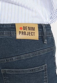 Denim jeans with a dark blue wash, featuring a tan leather label with "DENIM PROJECT" in black text. Includes back pocket.