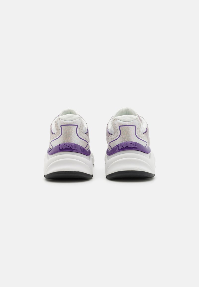 Zapatillas deportivas blancas con acentos morados, que cuentan con una parte superior texturizada, un collar acolchado y un detalle de logo en el talón. Suela de goma.