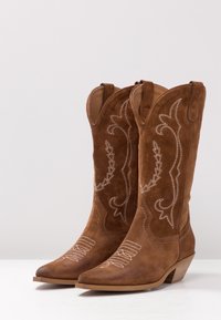 Brune cowboy-støvler i ruskind med spidse tæer, dekorativ hvid syning, en mellemhøj hæl og et traditionelt vestligt design.