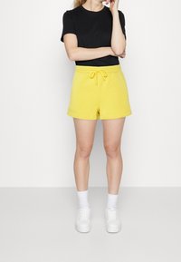 T-shirt noir à manches courtes associé à un short jaune vif avec une taille élastique et un cordon de serrage. Des baskets blanches et des chaussettes complètent la tenue.